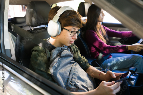 Fotografi Teenager wearing headphones and glasses, using a smartphone in a car while being