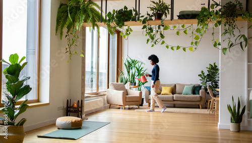 Authentic Black Woman walking, holding a watering can, during her mindful self-care routine in a bright, tranquil urban jungle living room apartment.