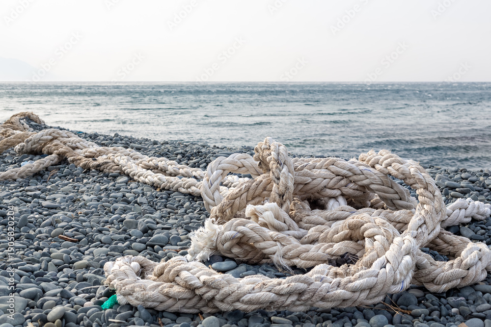 Fototapeta premium The ship's rope is in selective focus. Woven with thick bundles of threads, the ship's rope lies on a pebble beach. Türkiye, Samandağ, Hatay, Mediterranean coast.