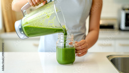 Close up of hands pouring green smoothie into glass jar in modern kitchen with natural light