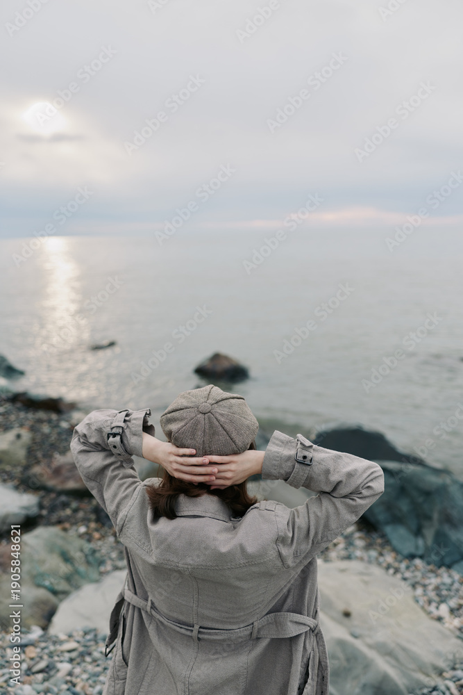 Obraz premium A person in a textured coat and knitted hat stands on rocky shore looking at calm sea under cloudy sky. Concept of solitude, nature, peaceful moment, and cold weather outdoors.