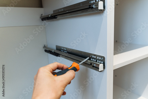 Closeup Screwdriver Adjusting Drawer Slide, Installer Tightening Rails On White Cabinet Interior With Visible Screws And Metal Hardware In Bright Home Setting, Focused DIY Assembly Atmosphere.