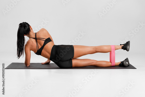 Fitness woman doing leg raises exercise for glute with resistance band on gray background. Athletic girl working out