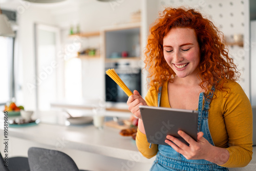 Fotografie Smiling young red haired woman cooking breakfast in bright modern kitchen using