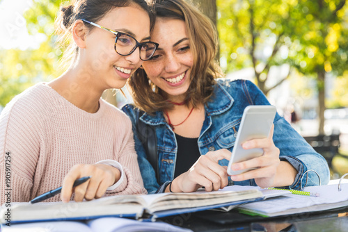 Two happy students sharing smartphone outdoors while studying