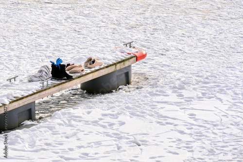 Winter boots, shoes, and clothing left on a snow-covered wooden dock next to a frozen body of water. The scene suggests a break from outdoor cold weather activities or a refreshing dip in icy water.