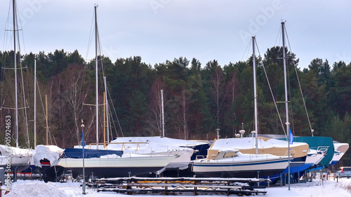 Several sailboats covered in snow are stored on the hard in a winter boatyard. The boats are parked on stands against a background of a forest line and a cloudy sky, illustrating seasonal storage.
