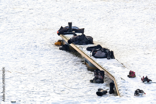 Various outdoor winter activity equipment, including ice skates, boots, and bags, left on a snowy wooden dock by a frozen lake. The scene depicts a break from winter sports 