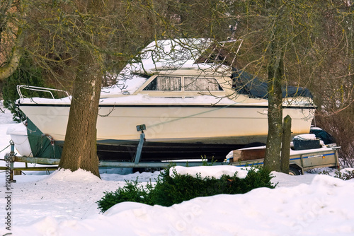 White cabin cruiser boat stored outdoors on a trailer covered in snow during winter. The boat is parked among bare trees and bushes on snowy ground, waiting for the spring season.