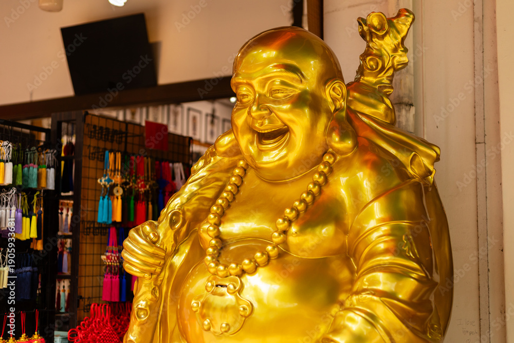 Fototapeta premium A large golden statue of a smiling figure stands in a shop filled with colorful items. Bright lights reflect off the shiny surface of the statue. This scene captures the vibrant energy of the market