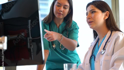 Two female medical professionals, a doctor and a nurse, collaborating and discussing patient data on a computer screen in a clinic office. The doctor takes a break to drink water from a mug.