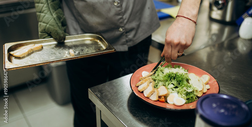 Close up of chef placing cooked chicken fillet onto plate while preparing salad. Professional restaurant kitchen food plating process.
