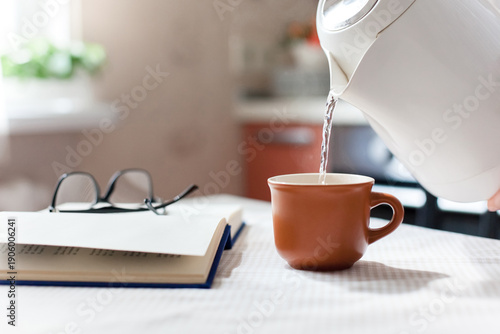 Morning tea at home in cozy kitchen. Slow living rituals, routine. Digital detox, mindfulness. Kettle pouring warm water. Cup of calming beverage, book, eyeglasses on table. Authentic lifestyle