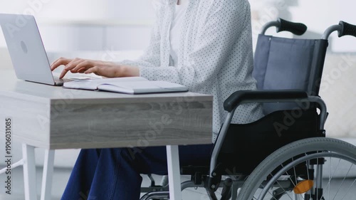 A person using a laptop sits at a desk in a well-lit room. The individual is in a wheelchair and focused on their work. Sunlight fills the space creating a bright atmosphere.