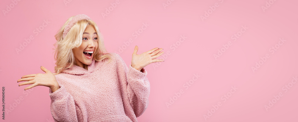 Fototapeta premium A girl with blonde hair is smiling widely while wearing a pink sweater. She has her hands raised in excitement. The background is a soft pink color.