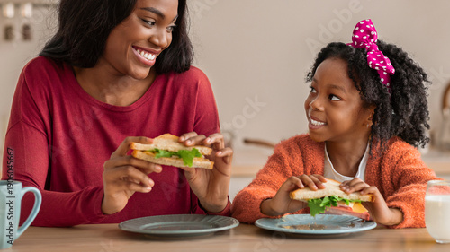 Tableau sur toile A mother and daughter sit at a kitchen table holding sandwiches while smiling at each other