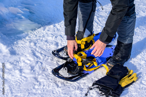 Close up of hands adjusting yellow straps on blue snowshoes resting on a snowy surface next to black and yellow gloves.
