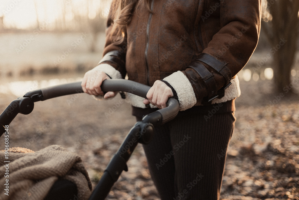 Fototapeta premium A woman in a brown aviator jacket holds her hands on the handle of a baby carriage while walking in the park at sunset