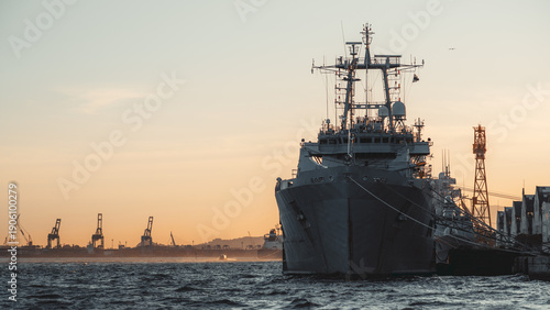 Front view of gray naval ship moored at industrial harbor at sunset, choppy sea foreground, port cranes and skyline in distance, warm golden sky with copy space