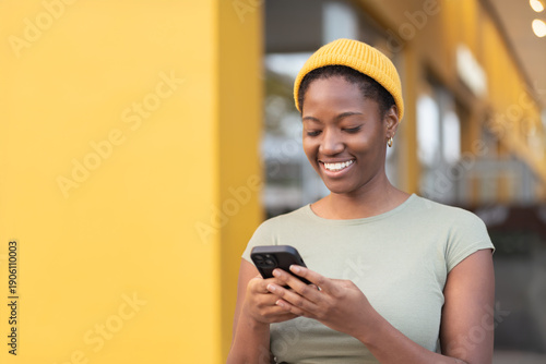 Smiling young black woman using smartphone against yellow wall. Happy female in yellow cap text messaging on phone during daytime