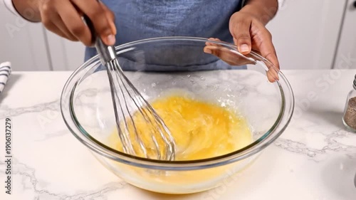 Hands whisking eggs in a clear glass bowl on a marble countertop