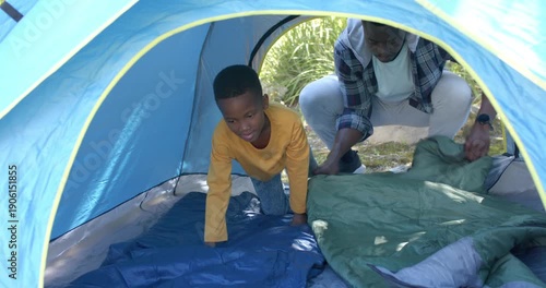 African-American father-son unfolding green bag and smoothing blue pad in blue tent preparing rest