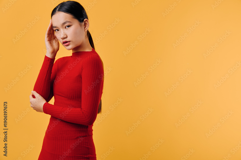 Obraz premium Young woman in red sweater with long sleeves poses thoughtfully against orange background. Model with long dark hair looks down with hand near head in studio shot.
