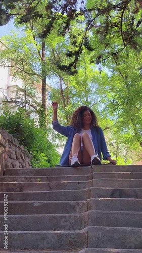 Happy young woman with curly hair dancing while sitting on a skateboard on stone stairs in a sunny park