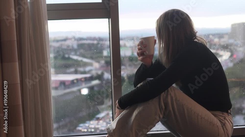 Young woman sitting on a windowsill and drinking coffee while looking at the city through a rainy window. Cozy home lifestyle scene with soft natural light, calm mood, urban view and quiet moment of