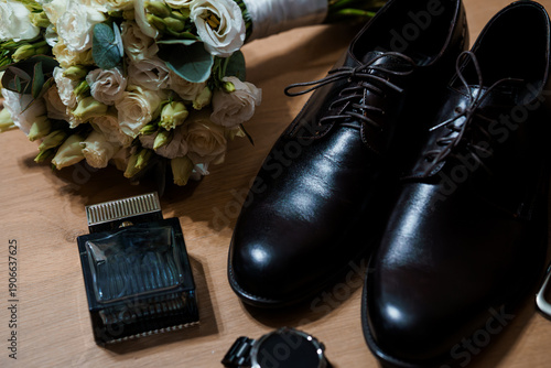 Elegant wedding groom's accessories with black dress shoes, perfume bottle, watch, and white floral bouquet on wooden surface