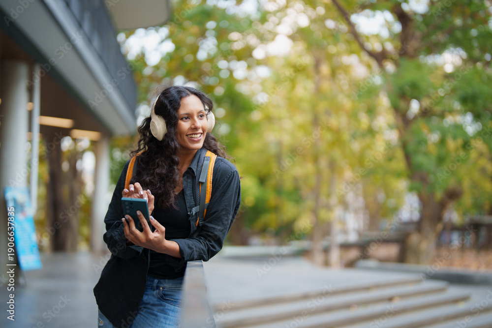 Fototapeta premium Young Hispanic college student with headphones smiling while listening to music on her smartphone outdoors on campus, casual backpack, enjoying a study break in autumn green surroundings