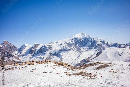 A wide panoramic view of the Himalayan mountain range featuring snow-covered peaks, rugged ridgelines, and a vast alpine landscape beneath a clear blue sky. The pristine winter terrain in Nepal