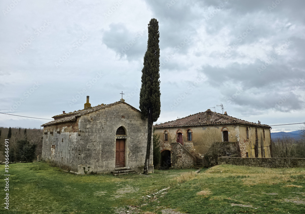Obraz premium This medieval Tuscan hamlet features an ancient stone church and historic buildings nestled in a rural landscape near a cypress tree