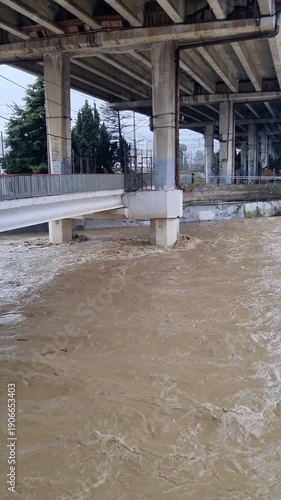 Raging muddy river flooding with powerful currents flowing under concrete bridge supports during a severe storm, highlighting climate change and natural disaster damage