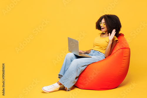 Fotografie Full body young IT woman she wear t-shirt casual clothes sit in bag chair hold use work on laptop pc computer waving hand get video call isolated on plain yellow orange background