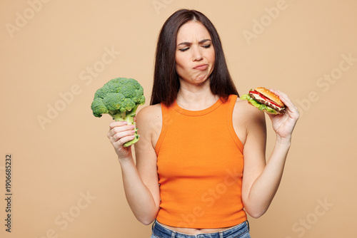 Bild auf Leinwand Young minded woman wear orange casual clothes hold eat choose between broccoli and burger isolated on plain pastel light beige background