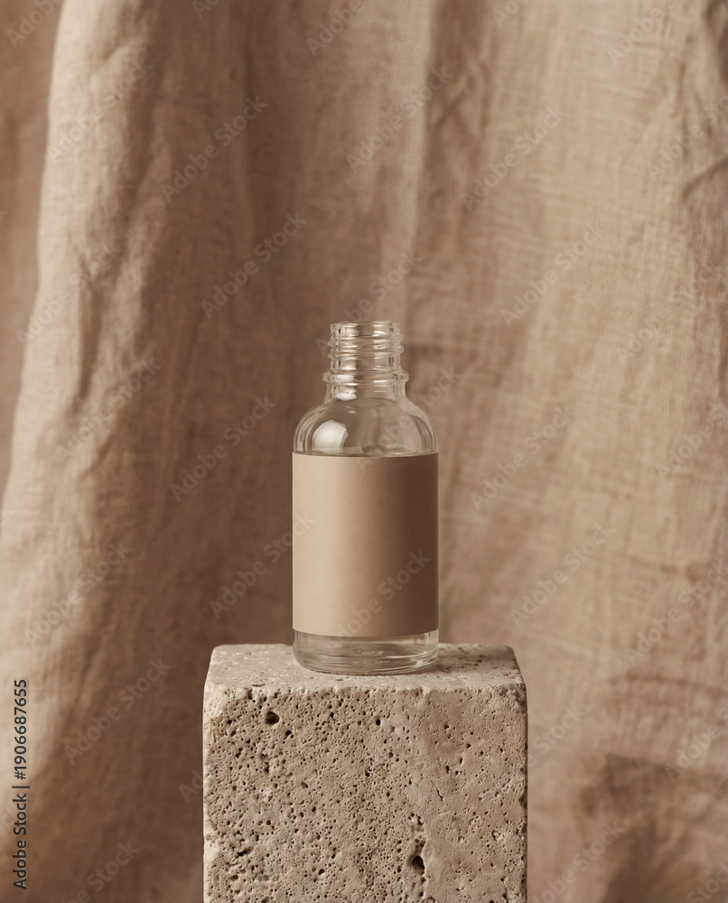 Naklejka premium A clear bottle with a blank beige label stands on a porous stone cube. The background consists of draped, textured beige fabric, creating a monochromatic and minimalist aesthetic.