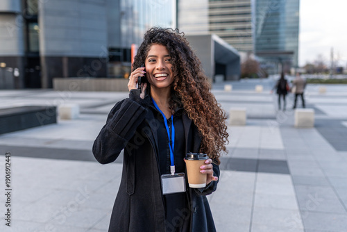 Young professional woman talking on phone, holding coffee cup against city buildings background, smiling