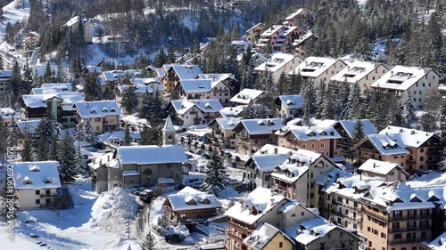 Aerial view of snow-blanketed buildings nestled among snow-covered trees, creating a striking contrast of white against the dark evergreens, Claviere, Piedmont, Italy.