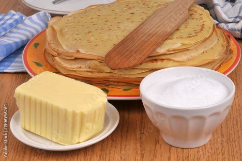 Morceau de beurre et ramequin de sucre en poudre devant une assiette de crêpes bretonnes avec une spatule en bois