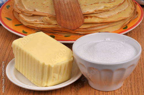 Morceau de beurre et ramequin de sucre en poudre devant une assiette de crêpes bretonnes avec une spatule en bois