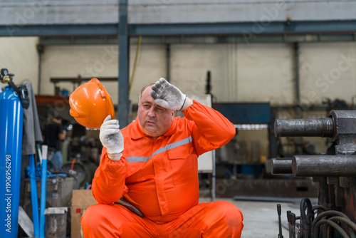 Exhausted industrial worker taking break wiping sweat from forehead after working hard in hot factory. Tired technician holding safety helmet feeling heat stroke during maintenance in workshop