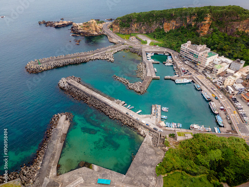Aerial view of Wanli Fishing Harbor in Taiwan, showing a small harbor sheltered by multiple concrete piers, surrounded by clear blue ocean water and a rugged coastal setting.

