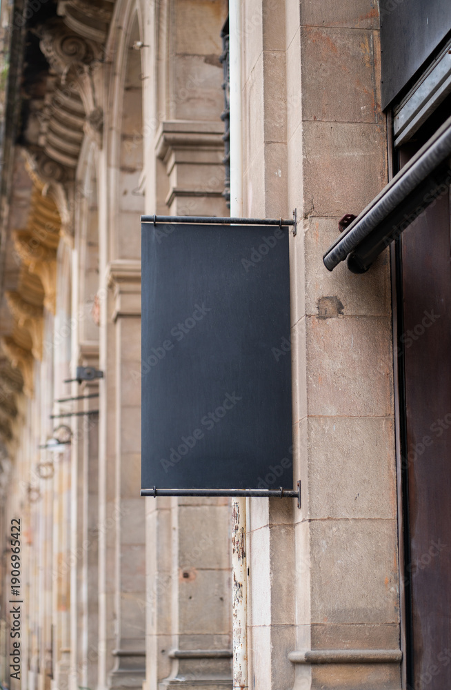 Fototapeta premium Blank black shop sign mockup hanging on historic stone wall, providing space for branding, advertising, and street navigation in urban area