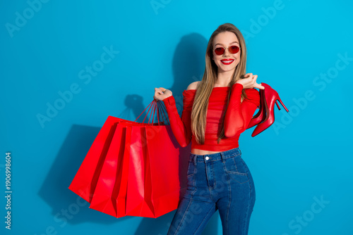 Stylish young woman in red outfit with shopping bags and heels against blue background showcasing glamour and shopping spirit