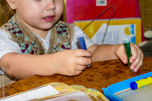 fair-haired little girl plays with decorative sparkles, holds in her hands, glitter in tubes, blue, green colors, view straight,  curls of hair, embroidered original vest, blondes