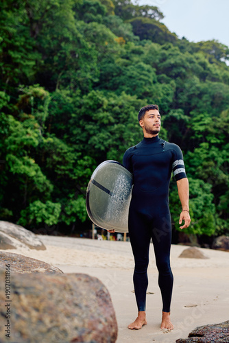 Konstfotografi Young man in a wetsuit standing on a sandy tropical beach with a surfboard, look
