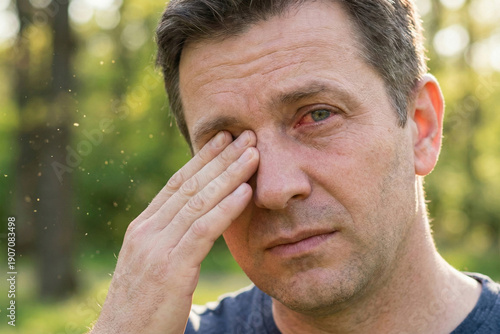 Mature man rubbing irritated itchy eye outdoors in park with visible pollen particles in air. Seasonal allergy and conjunctivitis concept.