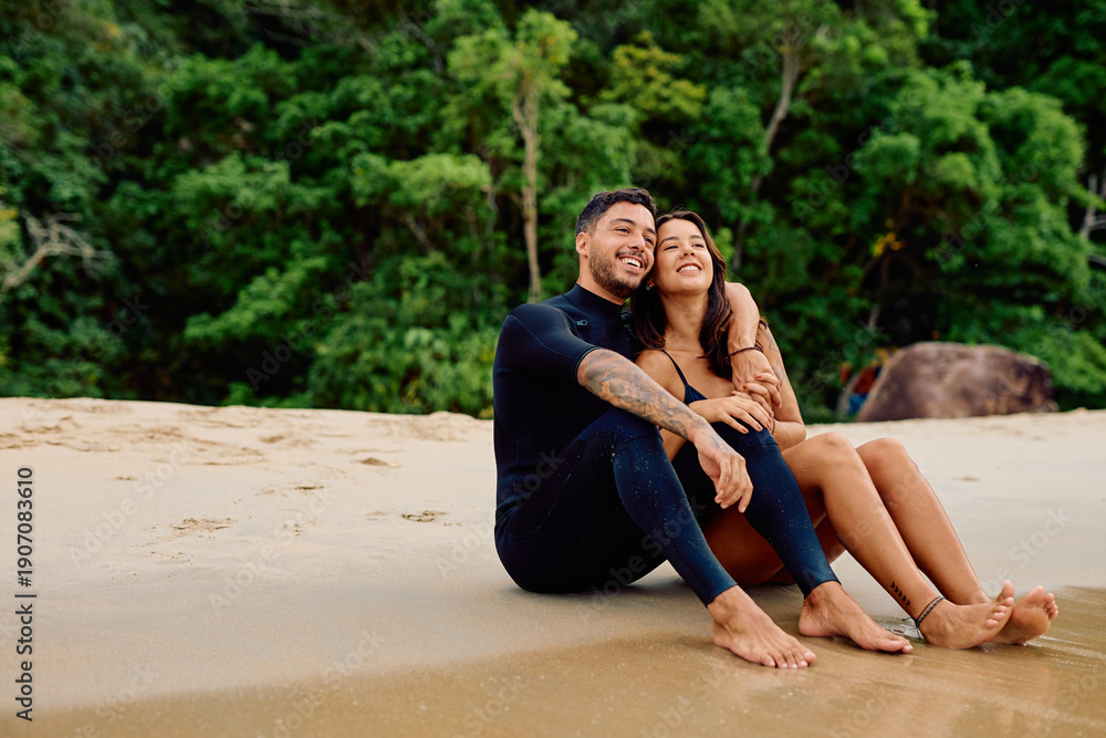 Fototapeta premium Smiling young couple in wetsuits sitting on a tropical beach, looking away while enjoying their summer holiday