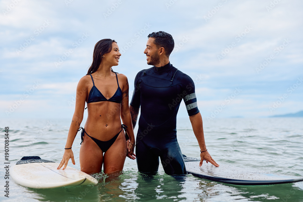Obraz premium Couple smiling and holding hands in ocean water with surfboards, preparing for activity. Concept of love, outdoor sport, and leisure